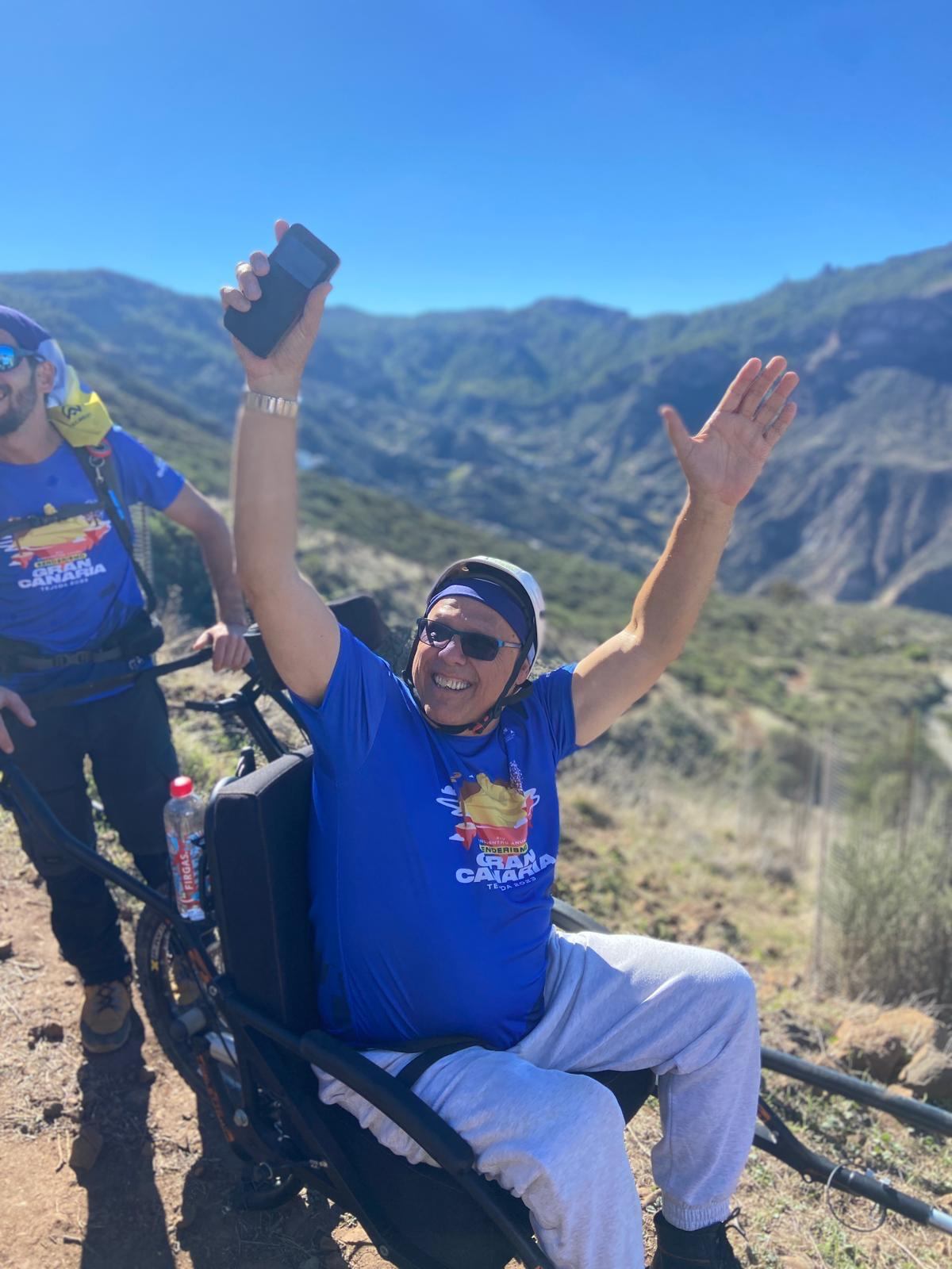 Participante en silla Joëlette celebrando con los brazos en alto frente al Roque Nublo